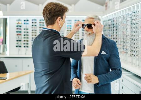 Vista laterale dell'ottico professionista in maschera che aiuta il maschio anziano mettere gli occhiali da sole mentre si lavora in un negozio ottico Foto Stock