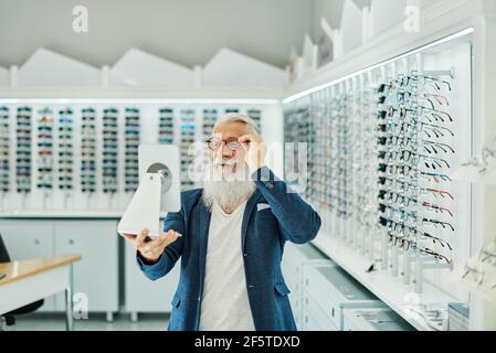 Soddisfare il maschio anziano elegante guardando nello specchio mentre provando sopra e scegliendo gli occhiali nel negozio ottico moderno Foto Stock