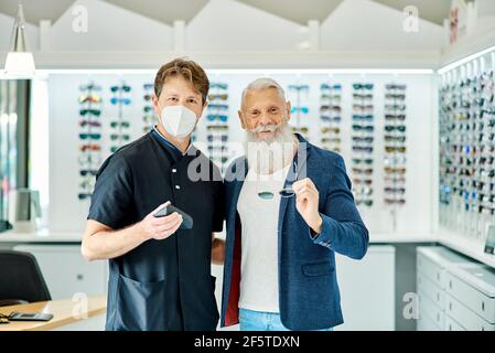 Sorridente cliente maschile anziano e ottico in piedi con gli occhiali dentro archiviazione ottica e vista della fotocamera Foto Stock