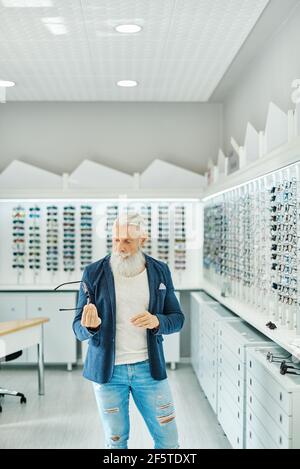 Anziani seri sopportato maschio in outfit trendy in piedi con moderno occhiali eleganti in un negozio di ottica Foto Stock