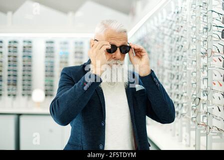 Senior maschile in outfit alla moda cercando su occhiali da sole di stile in moderno magazzino ottico Foto Stock