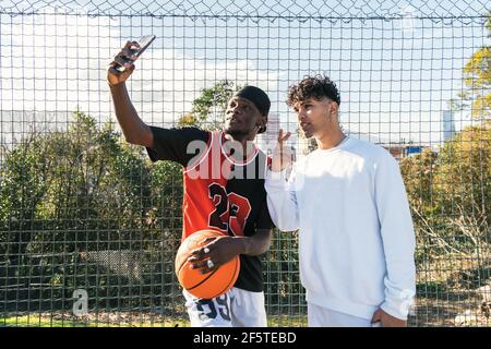 Giovani giocatori di streetball africani americani maschi in piedi sul campo da gioco di pallacanestro e portare selfie sullo smartphone Foto Stock