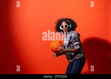 Contenuto giovane donna afroamericana in outfit e occhiali da sole alla moda ascoltare musica in cuffia e giocare con la palla da basket mentre si guarda Foto Stock