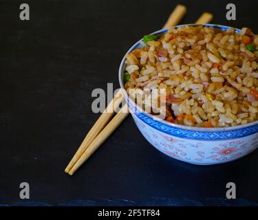 Riso fritto speciale in una ciotola di ceramica e bastoncini asiatici su tavola di ardesia nera Foto Stock