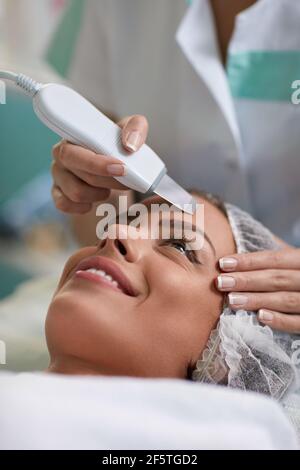 Primo piano di mani di cosmetologo con apparecchiature professionali a ultrasuoni per la pulizia della pelle sul viso della donna nel centro benessere Foto Stock