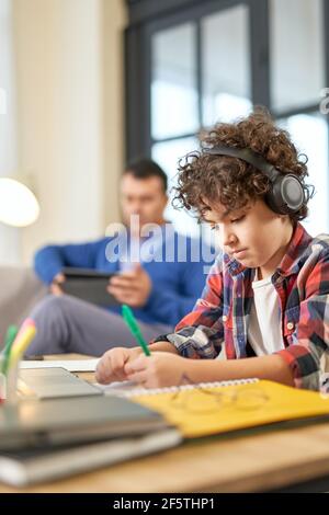 Entusiasta ragazzo della scuola latino-americana che indossa le cuffie, fa il lavoro scolastico e scrive sulla carta mentre si siede alla scrivania a casa Foto Stock