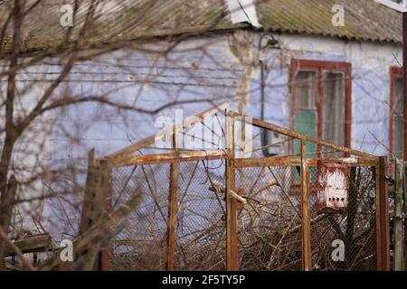 Vecchia casella postale arrugginita appesa su una rete di recinzione rurale. Casa blu su sfondo sfocato Foto Stock