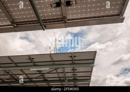 Linea elettrica aerea e torre di trasmissione vista da sotto una serie di pannelli solari. Foto Stock