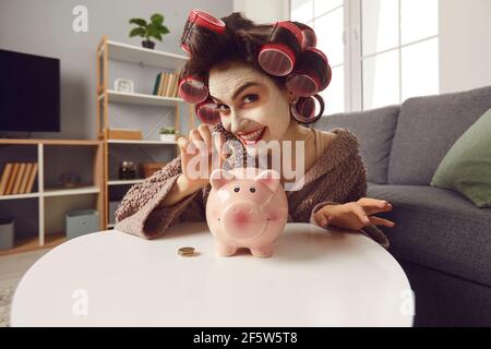 Giovane donna sorridente con maschera facciale e arricciacapelli monete nella sua banca piggy Foto Stock