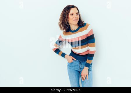 Bella giovane donna sicura che indossa felpa a righe casual e blu i jeans si levano in piedi tenendo la mano in tasca sorridendo i sguardi al lato su studio bianco Foto Stock