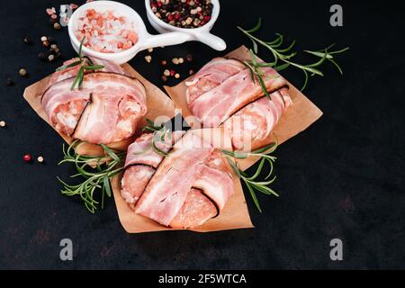 Tre hamburger di pollo avvolti in pancetta su carta da caverna con rami di rosmarino, pepe nero e sale rosa in piccoli piatti bianchi, fondo nero. Fa Foto Stock