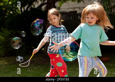 Due ragazze felici, bambini che giocano con grandi bolle di sapone all'esterno, bambini allegri nel giardino all'aperto divertendosi con le bolle che corrono intorno Foto Stock