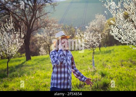 Sorridente agricoltore che parla con smartphone mentre esamina alberi da frutto fioriti in frutteto. Azienda agricola biologica di controllo a primavera Foto Stock