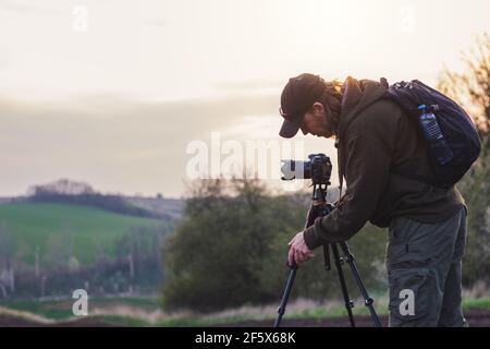 Fotografo di paesaggi che imposta il treppiede prima di scattare la foto. Uomo con macchina fotografica all'aperto durante il tramonto Foto Stock