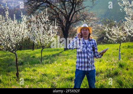 Agricoltore che utilizza smartphone e tablet mentre esamina frutteto fiorente. Giardiniere che parla con il telefono cellulare. Primavera in giardino Foto Stock