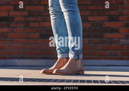Gambe femminili che indossano jeans e stivali in pelle in piedi accanto alla parete in mattoni. Donna che indossa abiti casual in città Foto Stock