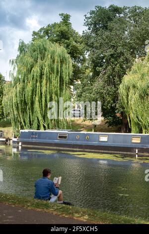 Uomo seduto da solo sulla riva del fiume che legge un libro vicino Il fiume Cam Cambridge Inghilterra Foto Stock