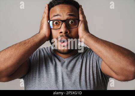 Asian unshaven uomo spaventato afferrando la testa e guardando telecamera isolata su sfondo grigio Foto Stock