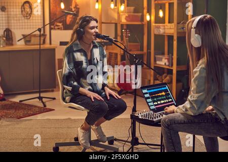 Giovane donna che lavora su un computer portatile con una giovane donna seduta di fronte lei e cantare in microfono in studio di registrazione Foto Stock