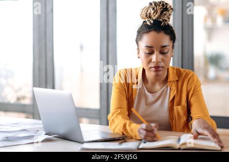 Focalizzato elegante ragazza afro-americana con dreadlock, studentessa, in abiti casual, studiare a casa usa un computer portatile, guardare lezioni online, prendere appunti in notebook, siede in soggiorno Foto Stock