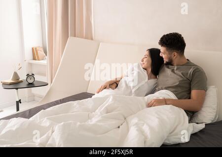 Giovane affettuosa coppia riposante che si rilassa su un grande letto matrimoniale la mattina e guardando attraverso la finestra mentre chiacchiera su piacevole roba Foto Stock