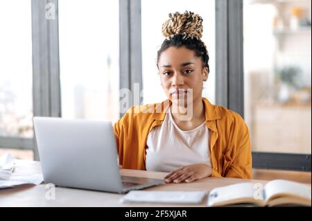 Ritratto di bella giovane donna afro-americana sicura con dreadlock, studentessa, in abiti casual di stile, con laptop, imparare a distanza, ascoltando una lezione online, guardare la fotocamera Foto Stock
