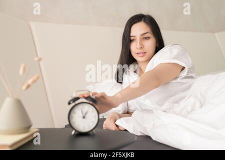 Giovane donna splendida in pigiami bianchi premendo il pulsante della sveglia mentre sedendo in su nel letto grande comodo dopo il sonno dentro la mattina Foto Stock