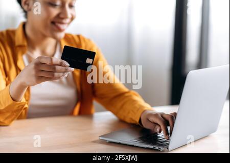 Donna afro-americana sfocata sorridente in abiti eleganti si siede a un computer portatile, paga per gli acquisti su Internet, digita i dati della carta di credito sulla tastiera, tiene la carta in mano. Shopping online Foto Stock