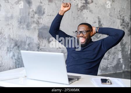 Emotivo giovane afroamericano che guarda lo schermo del portatile, festeggiando vincendo il tanto atteso gioco o ottenendo il prezzo in lotteria online, leggendo messaggio con incredibile piacevole notizia, è stato promosso Foto Stock