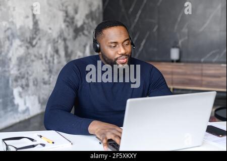 Uomo d'affari nero non rasato che fa la videoconferenza sul laptop seduto a casa, indossando la cuffia nera, la consulenza professionale dell'agente del call center, l'uomo che lavora in ufficio e parla, concetto di quarantena. Foto Stock