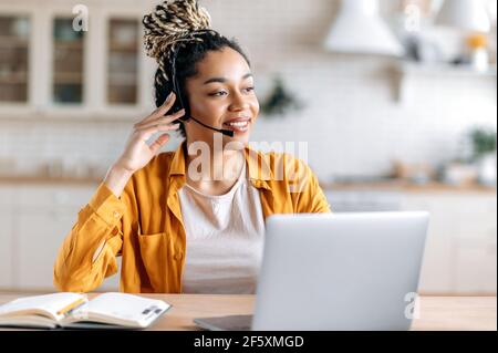 Gioiosa contented amichevole giovane afroamericana con dreadlock e cuffie, operatore di supporto, impiegato del call center, lavorando al portatile, consiglia i clienti online, guardando via, sorridendo Foto Stock