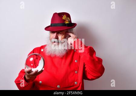 Vecchio maschio americano in una camicia rossa con un elegante cappello fedora che esamina con attenzione una palla di cristallo Foto Stock