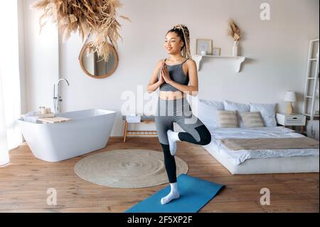Calma donna sportiva afroamericana con dreadlock, cura per la sua salute, fa yoga a casa su tappetino fitness, si erge in posa albero con le sue palme insieme, mantiene l'equilibrio, sorride Foto Stock