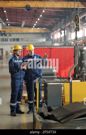 Due ingegneri si trovano in un'officina o in un impianto di grandi dimensioni e discutono nuove apparecchiature o macchine industriali mentre una di esse punta ad esso Foto Stock