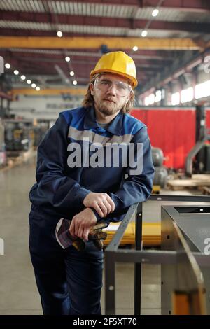 Giovane lavoratore di sesso maschile sopportato di grande pianta o fabbrica o. caposquadra in abbigliamento da lavoro e casco protettivo appoggiato alle apparecchiature industriali Foto Stock