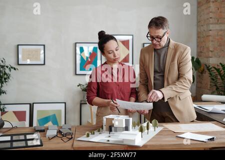 Giovane architetto femminile e il suo collega maschio maturo che tiene carta sopra il modello della nuova casa e del cantiere mentre discute i dettagli di schizzo Foto Stock