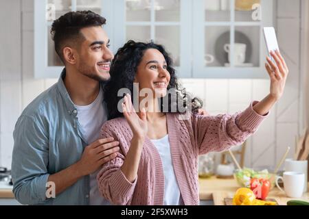 Riunione online. Allegra coppia mediorientale che utilizza lo smartphone in cucina per videochiamare Foto Stock