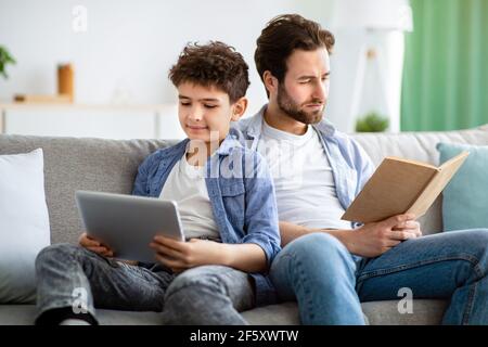 Nessuna comunicazione in famiglia. Padre leggere libro mentre suo figlio usa un tablet digitale, seduto insieme sul divano Foto Stock