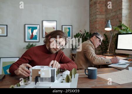 Giovane architetto maschile serio in occhiali che lavorano su modello di nuova casa e cortile contro il suo maturo disegno collega su carta Foto Stock