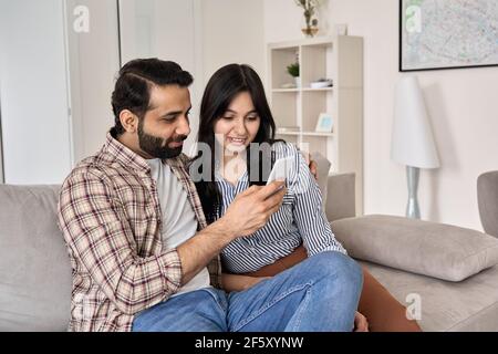 Felice giovane coppia indiana che usa il telefono seduto sul divano insieme a casa. Foto Stock
