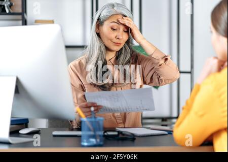 Una seccata scoraggiata stressata donna asiatica con capelli grigi maturi, donna d'affari, capo donna, in un ufficio moderno, insoddisfatto del lavoro, truffa un dipendente per un errore nei rapporti finanziari Foto Stock