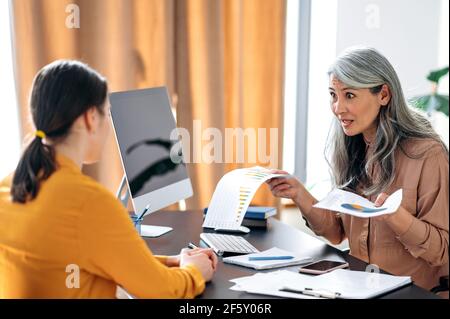 Arrabbiato indignante donna asiatica matura, business leader, capo, top manager, nel suo ufficio truffa un dipendente per un rapporto di affari mal redatto, gesti con le mani, tiene documenti Foto Stock