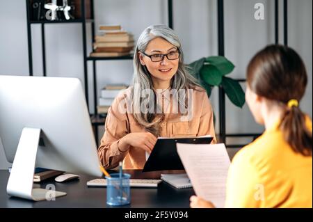 Intelligente fiducioso influente grigio-capelli asiatici donna indossare occhiali, business lady, manager HR, parlando con il candidato al lavoro in ufficio moderno, tiene e studi riprendere, sorridente amichevole Foto Stock