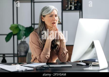 Scioccato grigio-capelli donna asiatica d'affari, freelancer, seduta al banco di lavoro e ricevere notizie inaspettate, messaggio, gesturando le mani mentre sorprende guardando lo schermo, stupito espressione sul suo volto Foto Stock