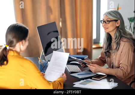 Donna asiatica intelligente e dai capelli grigi con occhiali, leader aziendale, manager HR, in ufficio moderno, parlando con la donna cercatrice di lavoro, tenendo e sfogliando il curriculum, sorridendo amichevole Foto Stock