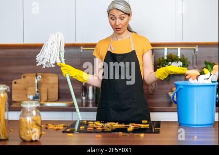 Indignato scioccato senior grigio-capelli casalinga asiatica o di pulizia lavoratore indossando guanti e grembiule, tenendo un mop, guardando il caos su stufa, gesturing con le mani, volto sorpreso Foto Stock