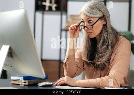 Scioccato entusiasta influente maturo grigio-capelli asiatici, donna d'affari, manager, è seduto al computer, ha ricevuto un messaggio inaspettato, guarda sorpreso lo schermo del computer, toglie gli occhiali Foto Stock