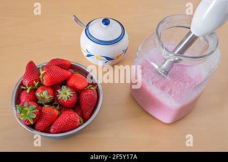 ciotola piena di fragole e una caraffa con frullato di fragola appena fatto, una ciotola di zucchero con un cucchiaio all'interno, un frullatore a immersione nella caraffa Foto Stock