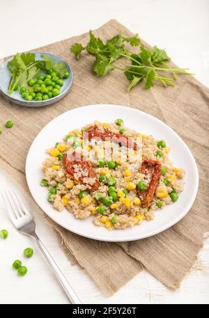 Porridge di quinoa con piselli verdi, mais e pomodori secchi su piastra di ceramica su fondo di legno bianco e tessuto di lino. Vista laterale, primo piano. Foto Stock