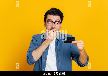 Scioccato giovane ragazzo caucasico intelligente con gli occhiali, sta coprendo la bocca con la mano, tiene e sorprende lo sguardo alla carta di credito, in piedi su sfondo arancione isolato, concetto senza contanti, spazio di copia, mockup Foto Stock